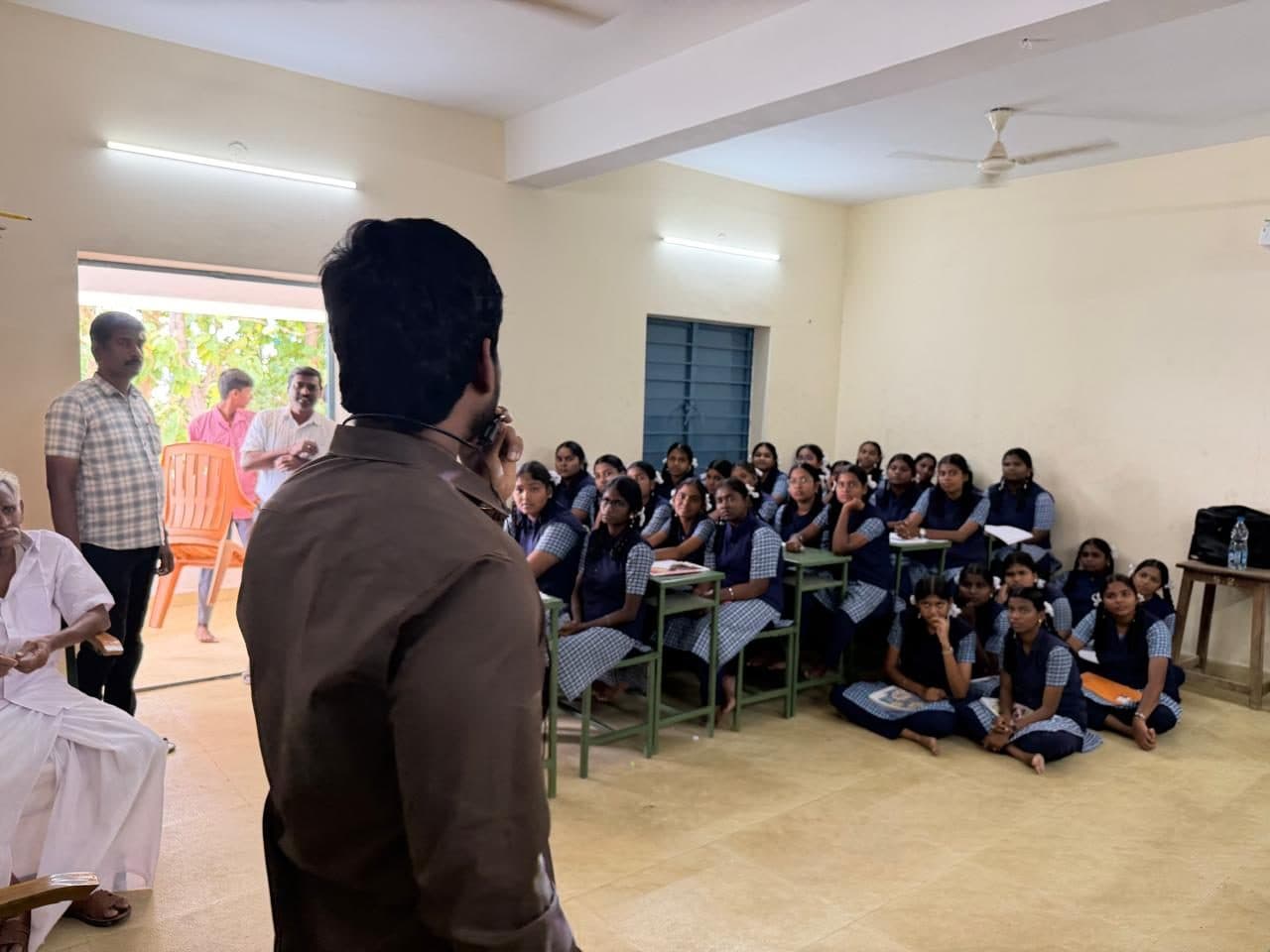 The full room seen from the front, students seated at desks and on the floor, teachers standing at the back.